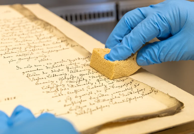 Das Bild zeigt zwei Hände mit Handschuhen über einer alten Handschrift. Eine Hand drückt einen Reinigungsschwamm auf das Archivgut.