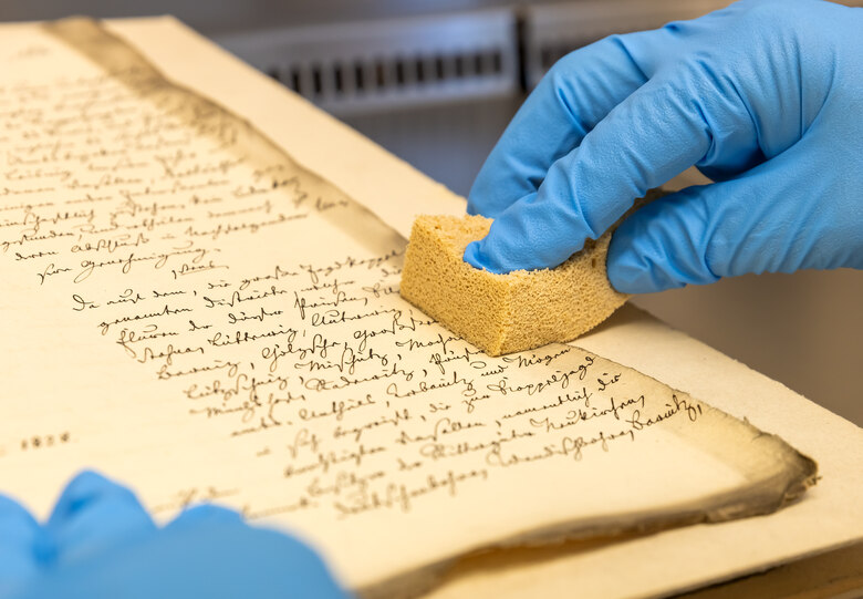 Das Bild zeigt zwei Hände mit Handschuhen über einer alten Handschrift. Eine Hand drückt einen Reinigungsschwamm auf das Archivgut.