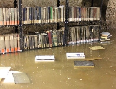 Regal mit Aktenbänden steht im hohen Wasser. Auf dem Wasser schwimmen Bücher.