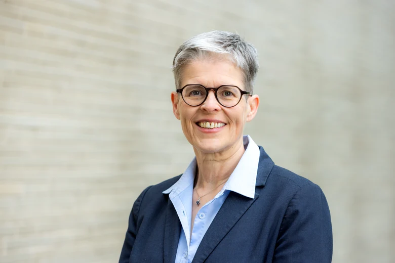 Das Foto zeigt eine Frau mit Brille vor einer Mauer stehend und lächelnd.