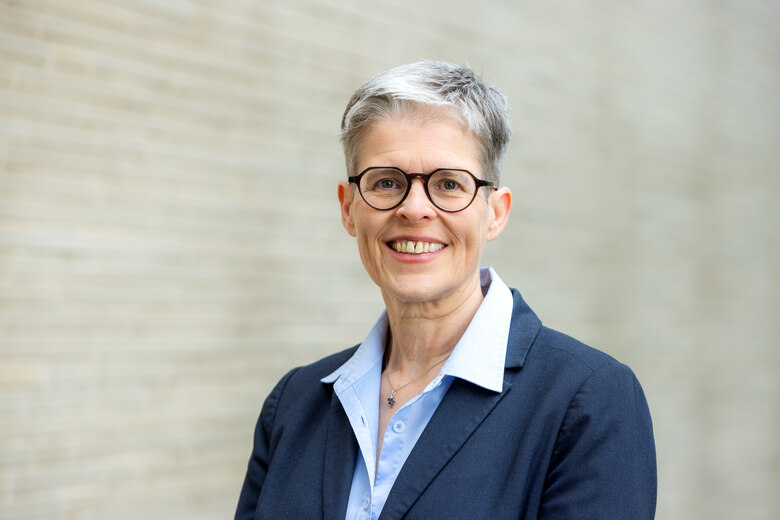 Das Foto zeigt eine Frau mit Brille vor einer Mauer stehend und lächelnd.